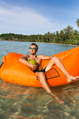 Young blonde man, boy at the beach with Lazy Inflatable Air Bed, hammock, couch camping.  Lounger Sofa Beach Chair Portable Sleeping Bag Mattress. Relax, vacation, travel. Coconut, sun glasses, hat