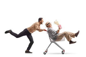 Mature man with a box of popcorn inside a shopping cart being pushed by a young man