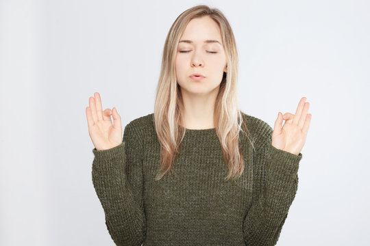 Yoga and meditation. Beautiful casually dressed young woman keeping eyes closed while meditating, exhaling, feeling relaxed, calm and peaceful after hard working day, holding hands in mudra sign.