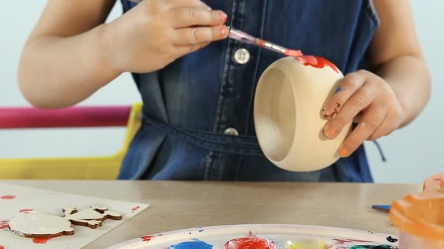 Child, 4-5 year old girl, in the studio. She paints and paints wooden toys. On the table, a palette for mixing paints. Girl in a denim dress. She has blond hair.