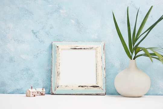 Square blue Photo frame mock up with green tropical plants in vaseand small wooden houses on shelf. Scandinavian style.  Text space