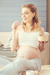 Sweeten your life. Cheerful pregnant woman eating macaroons and resting on the floor at home