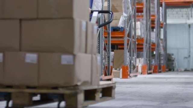 PAN of unrecognizable female worker wearing hard hat pulling platform cart with cardboard boxes through aisle with shelves in factory warehouse