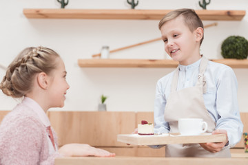 little waiter holding wooden tray in hands with dessert against kid sitting at table at cafe