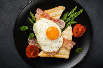 Breakfast or lunch with Fried egg, bread toast, green asparagus, tomatoes and bacon on black plate. Top view.