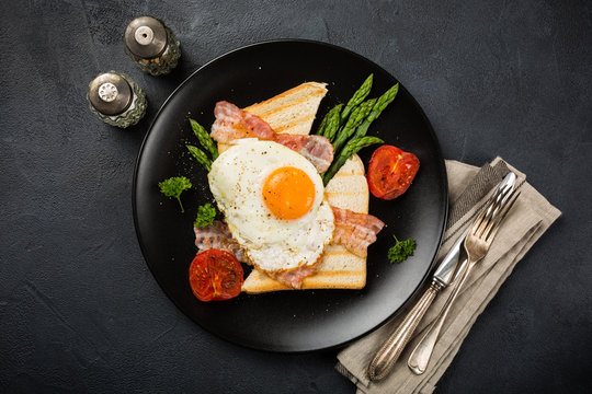 Overhead shot of breakfast or lunch with fried egg, bread toast, green asparagus, tomatoes and bacon on black plate with copy space.