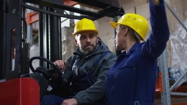 Slowmo of serious female warehouse manager in blue uniform and hard hat holding sheet of paper and explaining something to bearded male forklift operator
