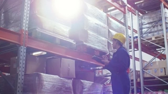 PAN of concentrated female worker in uniform and hard hat inspecting cardboard boxes on rack shelves and typing on tablet