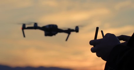 Close view of drone pilot's hands as he controls quadcopter hovering then flying off in the background. Aircraft and pilot silhouetted against sunset sky. Slow motion recorded at 60fps, 4K. - Powered by Adobe