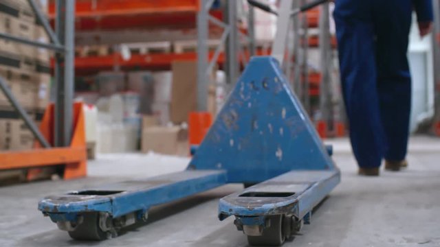 Tracking with low-section of unrecognizable worker in blue uniform pulling empty platform cart in warehouse