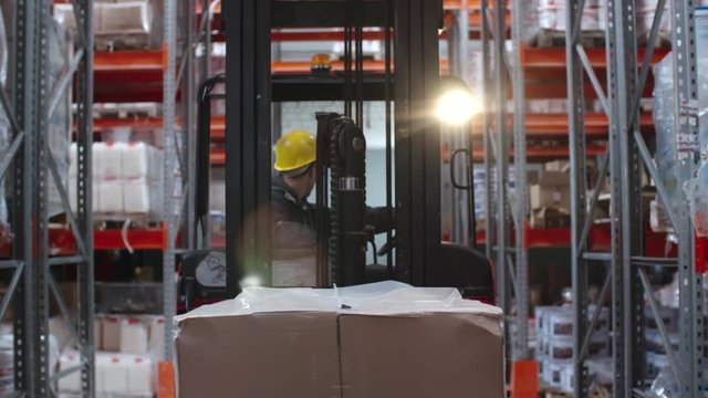 Tracking of male forklift operator in hard hat transporting cardboard box and driving between rack shelves in warehouse