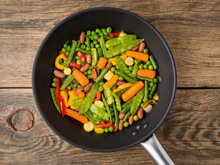 big pan fried with colorful vegetables - peppers, peas, green beans, baby corn, carrots, beans. Colorful vegetarian lunch, top view, wooden background,