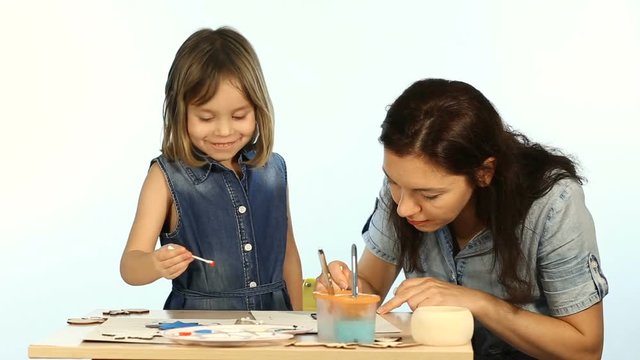 Shooting in the studio on a white background. A child, a funny girl, learns to draw. Mom shows you how to paint wooden New Year and Christmas figures. They are wearing a blue denim dress.