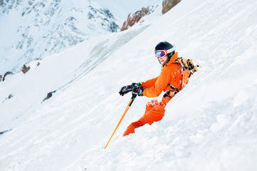 Fototapeta na wymiar A skier in an orange overall and with a backpack is sitting happy in the snow after falling.