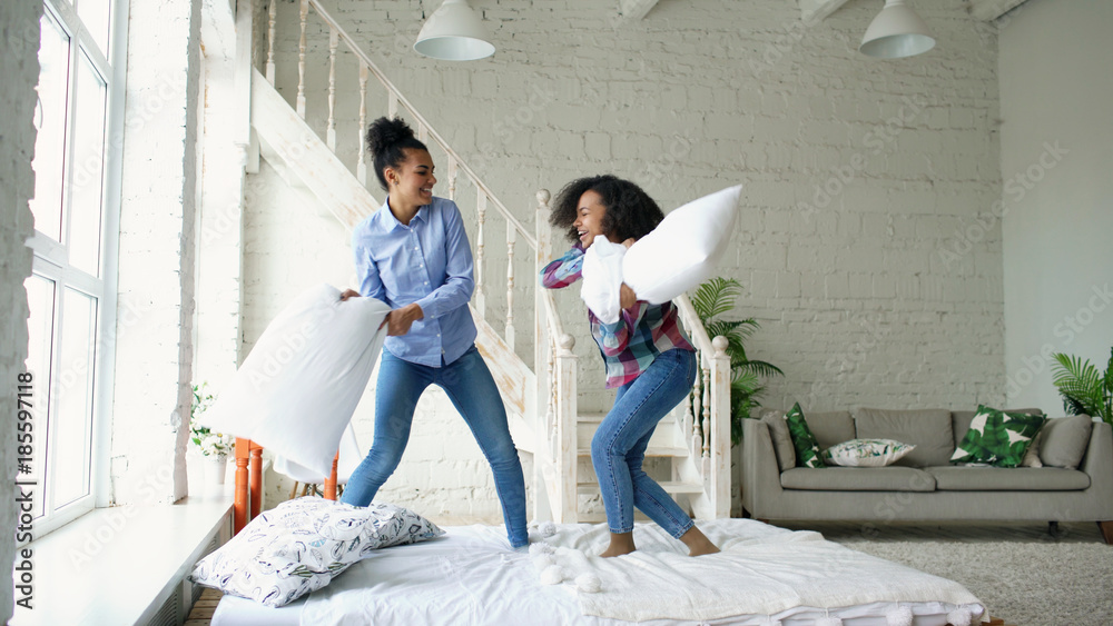 Wall mural Mixed race young pretty girls jumping on bed and fight pillows having fun at home