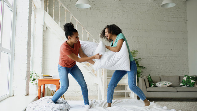 Mixed race young pretty girls jumping on bed and fight pillows having fun at home