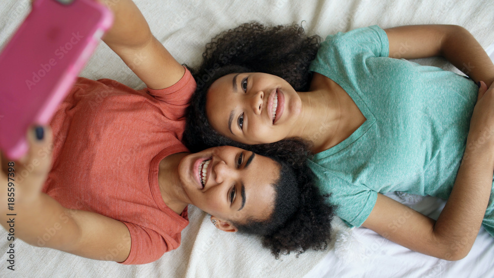 Wall mural Top view of beautiful mixed race funny girlfriends making selfie portrait on bed in bedroom at home