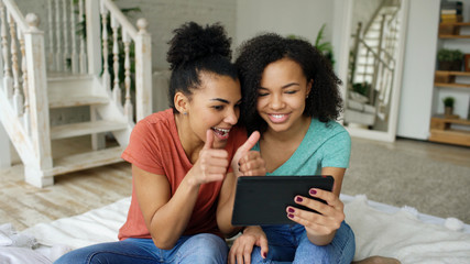Cheerful mixed race young funny girls talking online video chat on tablet computer with their friends at home