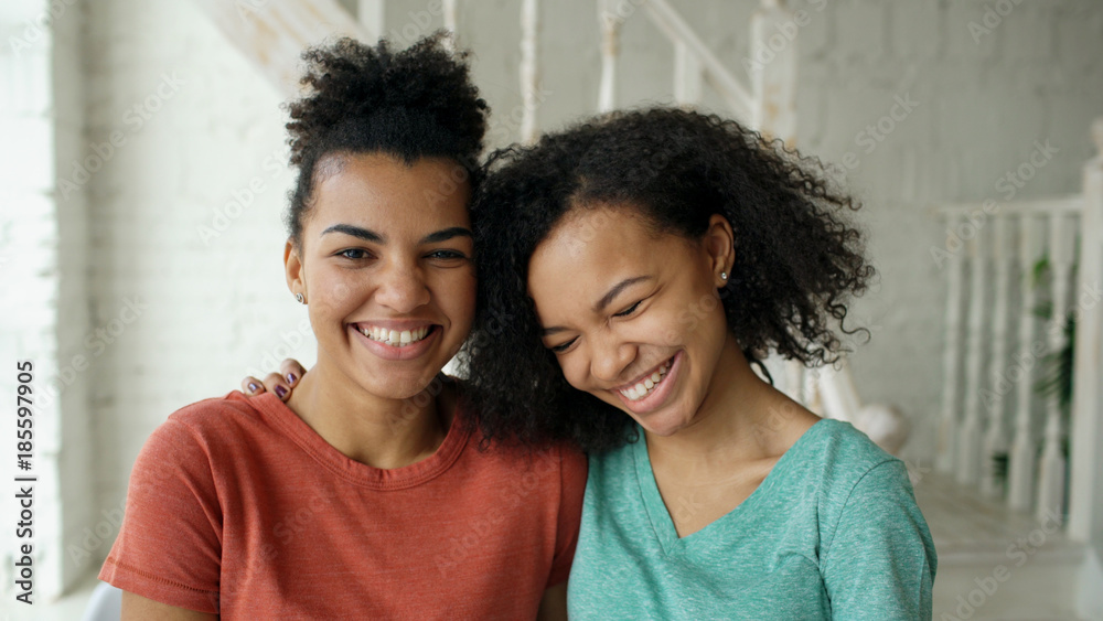 Wall mural Portrait of two beautiful african american girls laughing and looking into camera at home