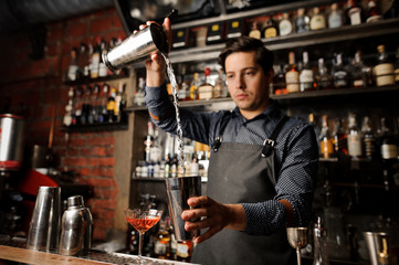 Young barman pouring alcoholic drink from one metal glass into another