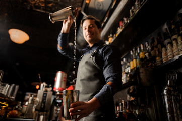 Fototapeta na wymiar Smiling barman pouring alcoholic drink from one metal glass into another