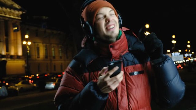 Young Caucasian man listening to music on headphones on your phone, dancing and singing for joy, eyes closed, enjoying the music in town tonight walking in the winter.