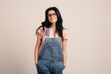 Beautiful smiling brunette girl wearing glasses in denim overall posing beside blank wall.