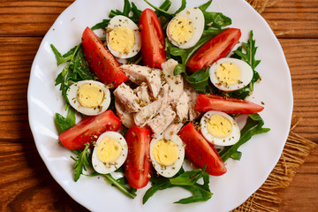 Fresh vegetable salad with boiled chicken breast and quail eggs. Salad with fresh tomatoes, rucola, quail eggs, chicken breast and spices on a serving plate and a wooden background. Closeup. Top view