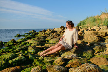 dark-haired girl in a white dress sitting on the rocks by the sea.