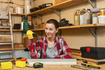 Beautiful caucasian young brown-hair woman in plaid shirt and gray T-shirt working in carpentry workshop at table place, drilling with drill holes in piece of iron and wood while making furniture.