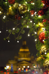 Fototapeta na wymiar Christmas baubles on fir branches and glowing garland outdoors in the evening, church on background / Decorations for the New Year and holidays in St. Petersburg on St. Isaac's Square