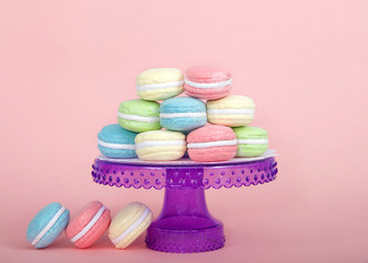 Large Macaroon cookies covered in granulated sugar on a table alternating colors stacked on a purple pedestal. Three cookies on the table. pink background. Popular pastry in France.