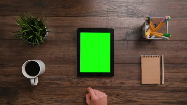 A man's finger scrolling on the green touchscreen. The i-Pad is on the brown table. View from the top. Close-up.