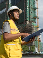 Medium vertical profile view of Asian engineer with hardhat and clipboard & building project in background