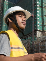 MCU Asian engineer with hardhat seen in 3/4 view, with construction in background