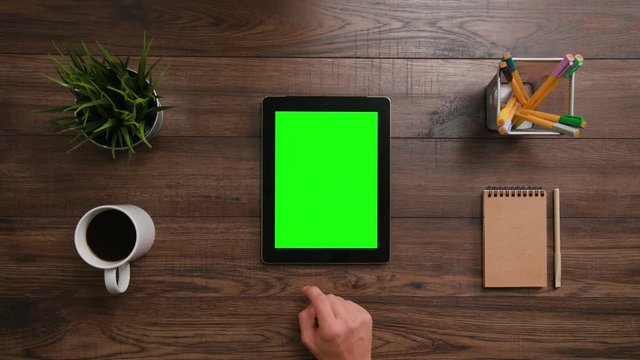A man's finger scrolling on the green touchscreen. The i-Pad is on the brown table. View from the top. Close-up.