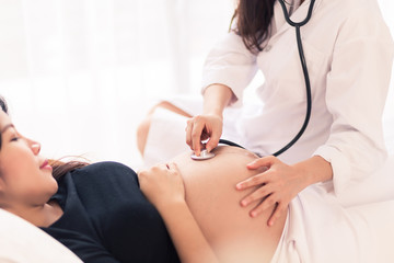 Female Obstetrician doctor with stethoscope listening to pregnant woman baby heartbeat at hospital.