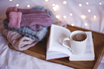 Cup of coffee staying on open book with stack of knitted clothes: gloves, hat, scarf on wooden tray over lights at background. Winter season. Cozy atmosphere.