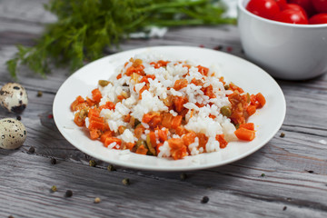 delicious rice with stewed carrots, peas and onions on a white plate, quail eggs, bowl with cherry tomatoes, towel, pepper and dill on a light wooden table