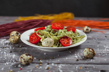 pasta from green spinach with mozzarella, greens and French mustard, quail eggs and cherry tomatoes on a plate, bell pepper and large sea salt on a gray wooden table on a dark background