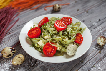 pasta from green spinach with mozzarella, greens and French mustard, quail eggs and cherry tomatoes on a plate, bell pepper and large sea salt on a gray wooden table on a dark background