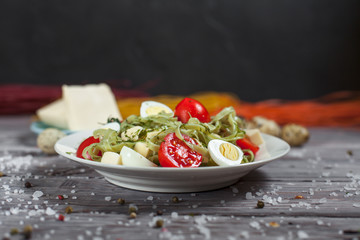 pasta from green spinach with mozzarella, greens and French mustard, quail eggs and cherry tomatoes on a plate, bell pepper and large sea salt on a gray wooden table on a dark background