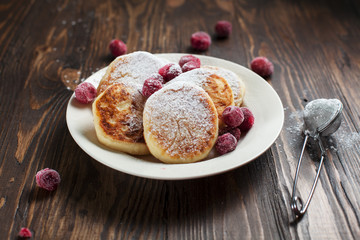 syrniki with powdered sugar on a white plate, sieve and frozen berries on a dark wooden table