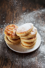syrniki with powdered sugar on a white plate, a sieve and caramel topping on a dark wooden table