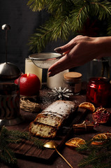 christmas cake, in germany christstollen, woman hand sprinkling powdered sugar, dark background