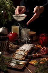 christmas cake, in germany christstollen, woman hand sprinkling powdered sugar, dark background