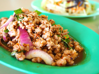 Close up Thai local traditional Isaan Northest Larb Moo, raw semi stir fried spicy minced pork with shallot, spring onion, and herb, on green oval plastic plate, with blurred Som Tam, papaya salad