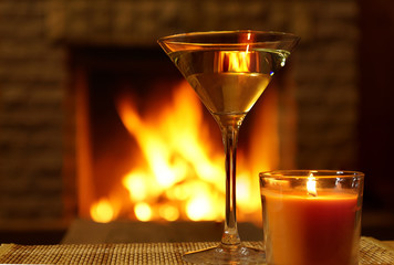 Martini in wineglass and candle over burning cozy fireplace background.