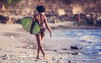 Surfer girl with afro hairstyle walking with surfboard on the Uluwatu beach at sunset in Bali, Indonesia