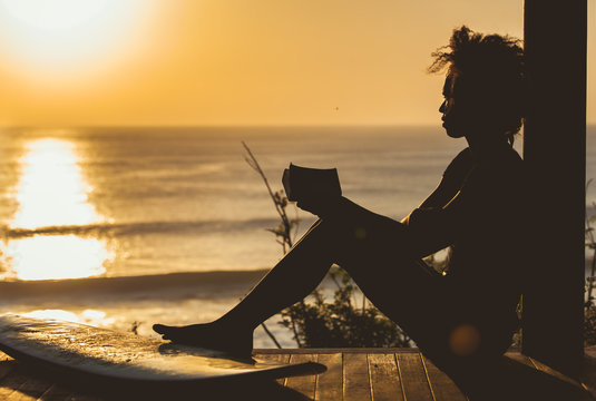 Beautiful surfer girl with afro hair style reading book on top of the cliff at sunset on the beach in front of seascape in Bali, Indonesia. copy space